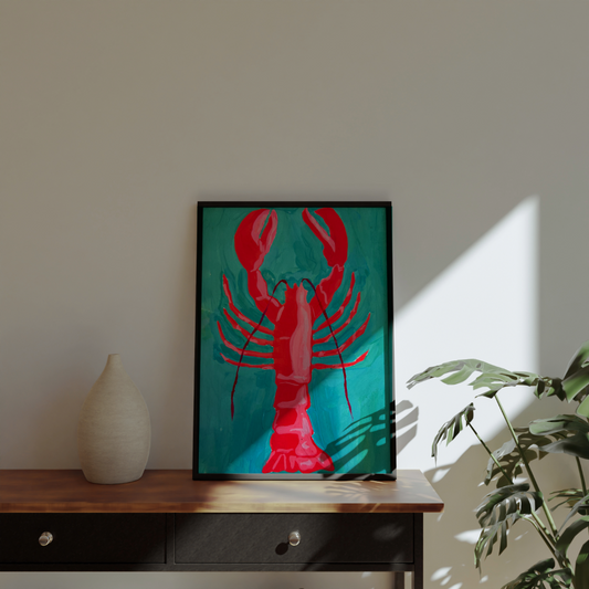 Framed artwork of a red lobster on a teal background, displayed on a wooden surface with a plant and vase in the background.