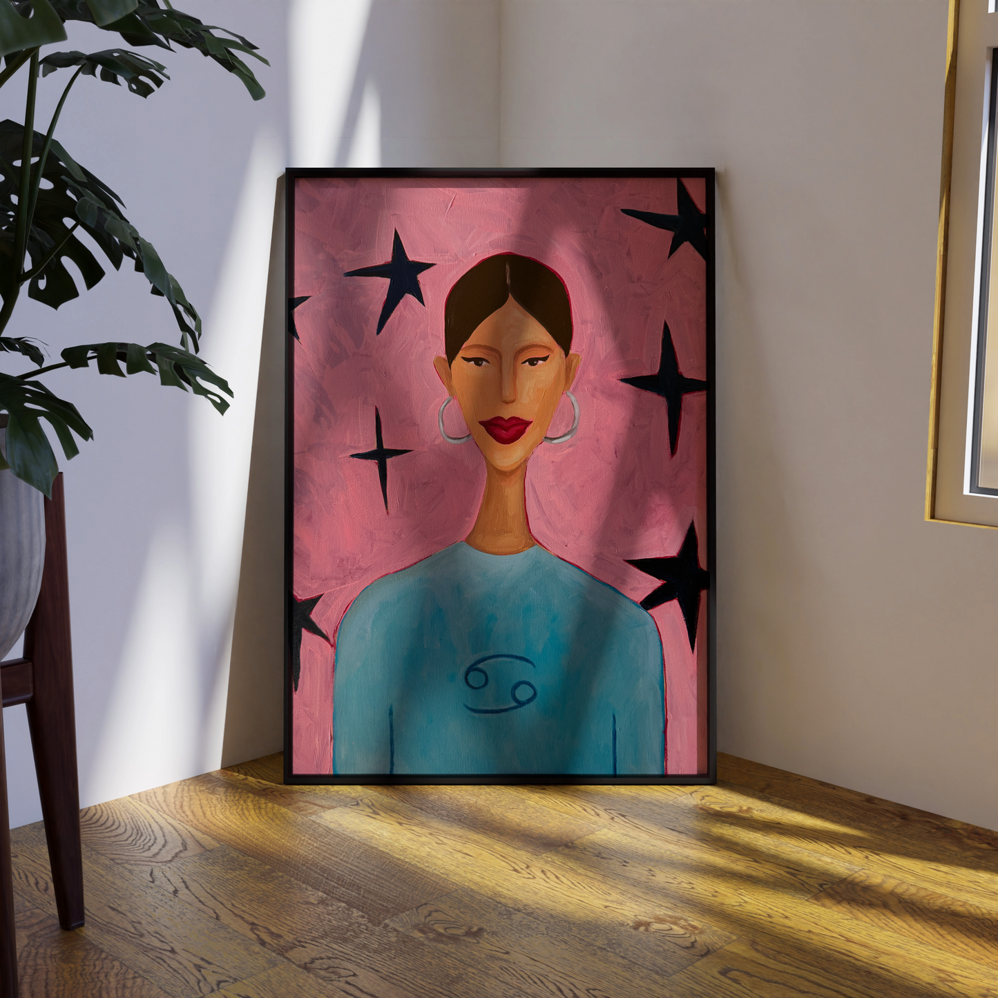 A framed art print depicting a woman with brown hair, red lips, and a blue top against a pink background with black stars, leaning against a wall on a wooden floor next to a potted plant.