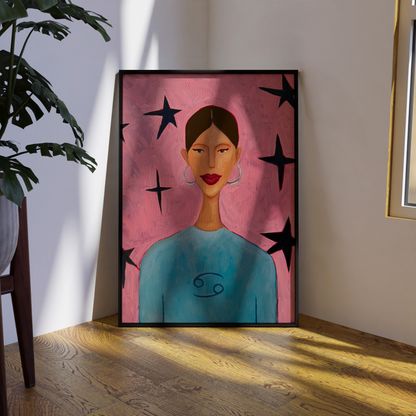A framed art print depicting a woman with brown hair, red lips, and a blue top against a pink background with black stars, leaning against a wall on a wooden floor next to a potted plant.